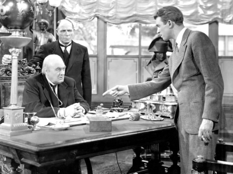 Henry Potter sits at his banker's desk, as George Bailey gestures angrily at him.