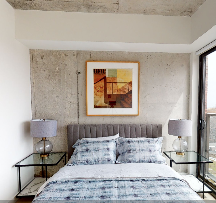 a bedroom with a concrete wall, bedside table, and warm colored painting above the bed