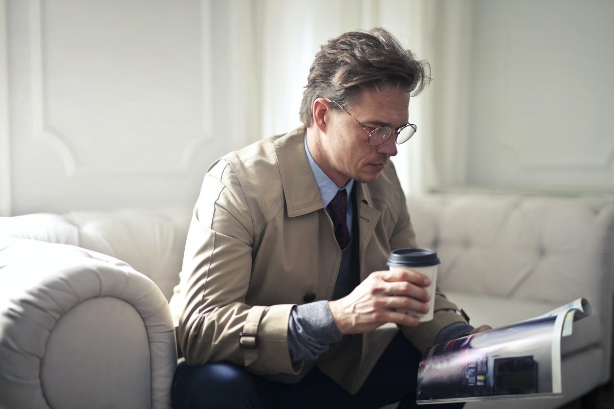 A middle aged man with a takeaway coffee sits on a beige leather couch and reads a magazine calmly.

