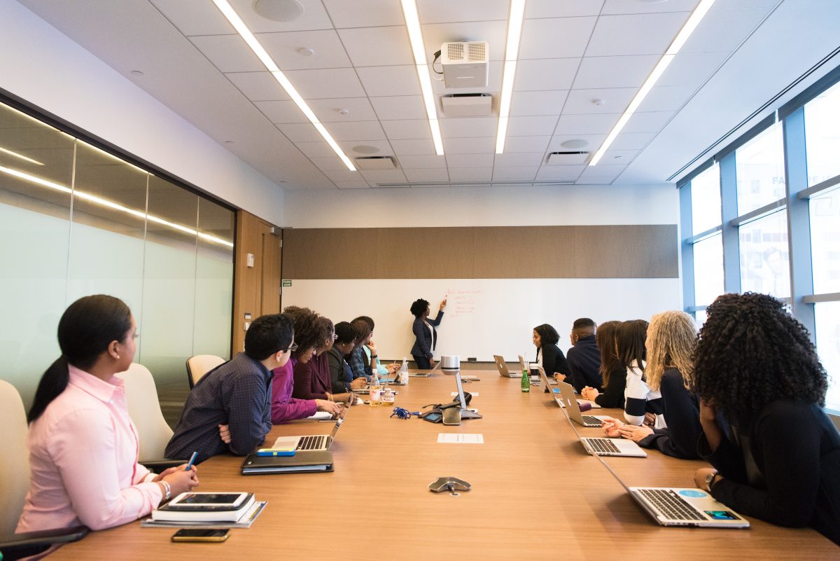 A group of people sit around a long conference table while someone gives a presentation. Bright fluorescent lights hang on the ceiling above.
