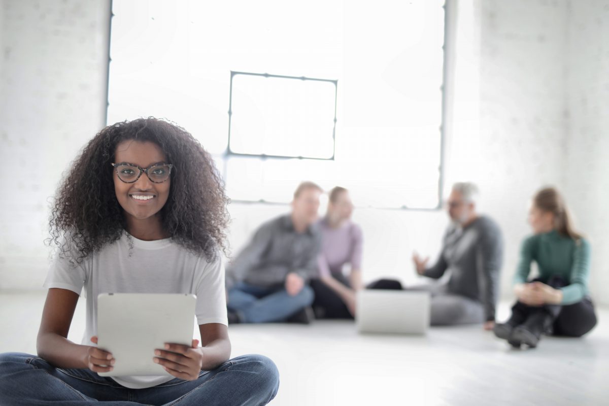  Une jeune femme noire portant des lunettes sourit et tient un iPad. En arrière-plan se trouve un groupe de personnes assises sur le sol rassemblées autour d'un ordinateur portable et discutant.