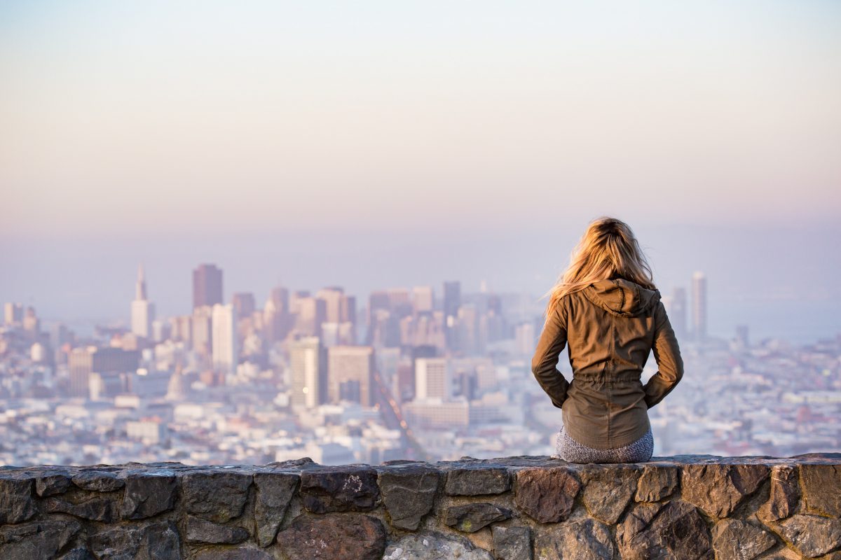 A woman on a rock platform views the city from far away. She faces away from the camera.